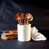 Several hand carved leaf shaped wooden salad servers in a white utensil crock on a black background
