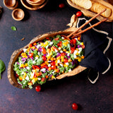 extra large rustic wood bowl, filled with colorful salad. Has salad servers sticking out of the bowl, along with a black napkin. All on a dark background with additional olive wood serving dishes in the background.