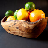 A carved wooden bowl with a natural live edge, filled with citrus fruits including oranges, limes, and a yellow citrus fruit, set against a dark background.