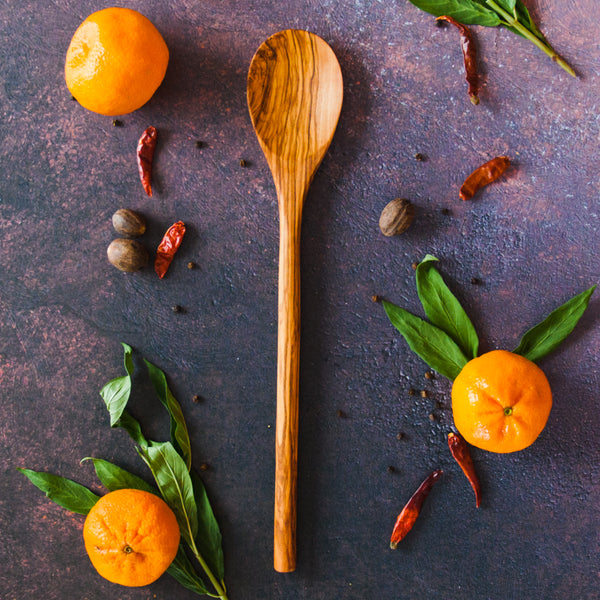 Olive wood kitchen spoon on a dark rusty background with clementines, green leaves and red chili peppers around it