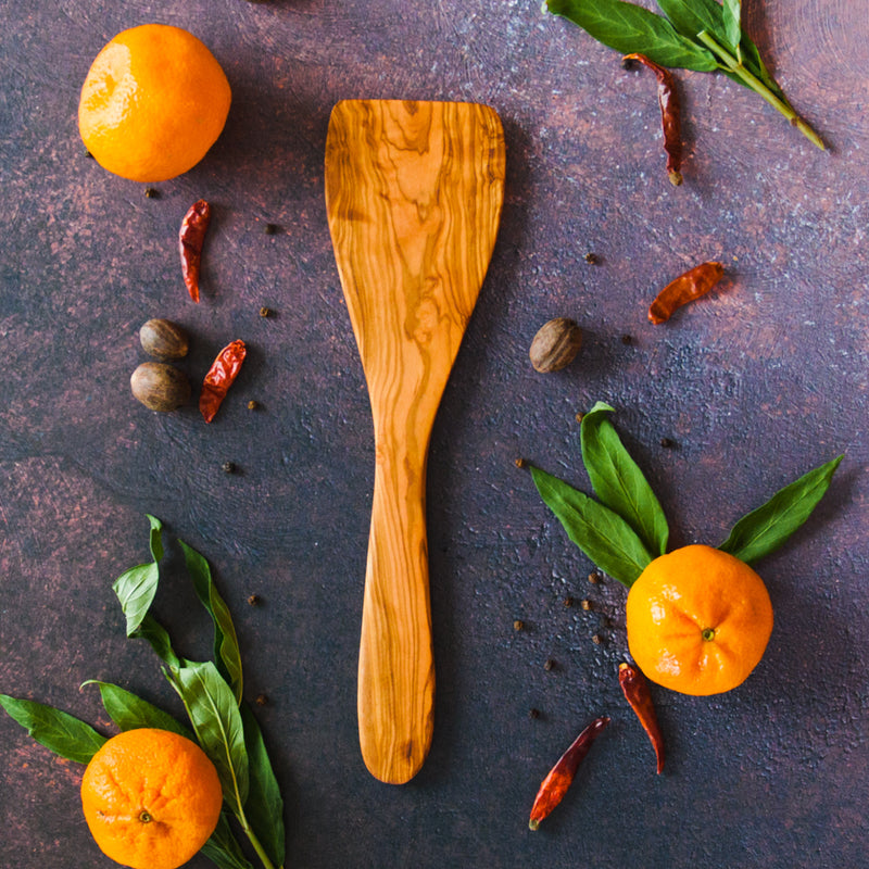 Olive wood spatula on a dark rusty surface with clementines, chilis and green leaves