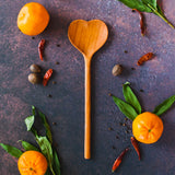 Large wooden heart shaped spoon on a dark rusty background with oranges, greenery and red peppers surrounding it