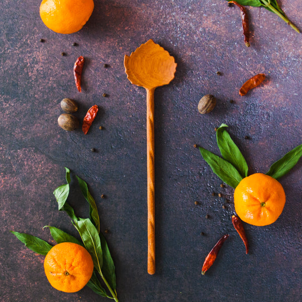 Aspen Leaf shaped wooden spoon on a dark background with oranges, green leaves and red chili peppers