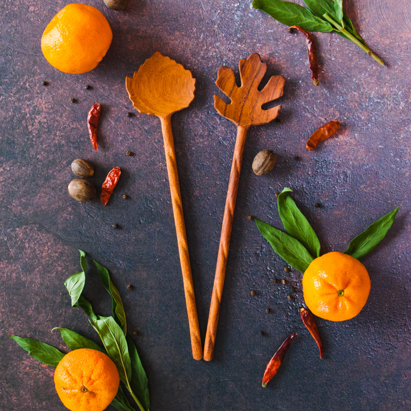 Two Wooden salad servers, shaped like leaves, with long twisted handles, all on a rusty dark background with oranges, greenery and red peppers