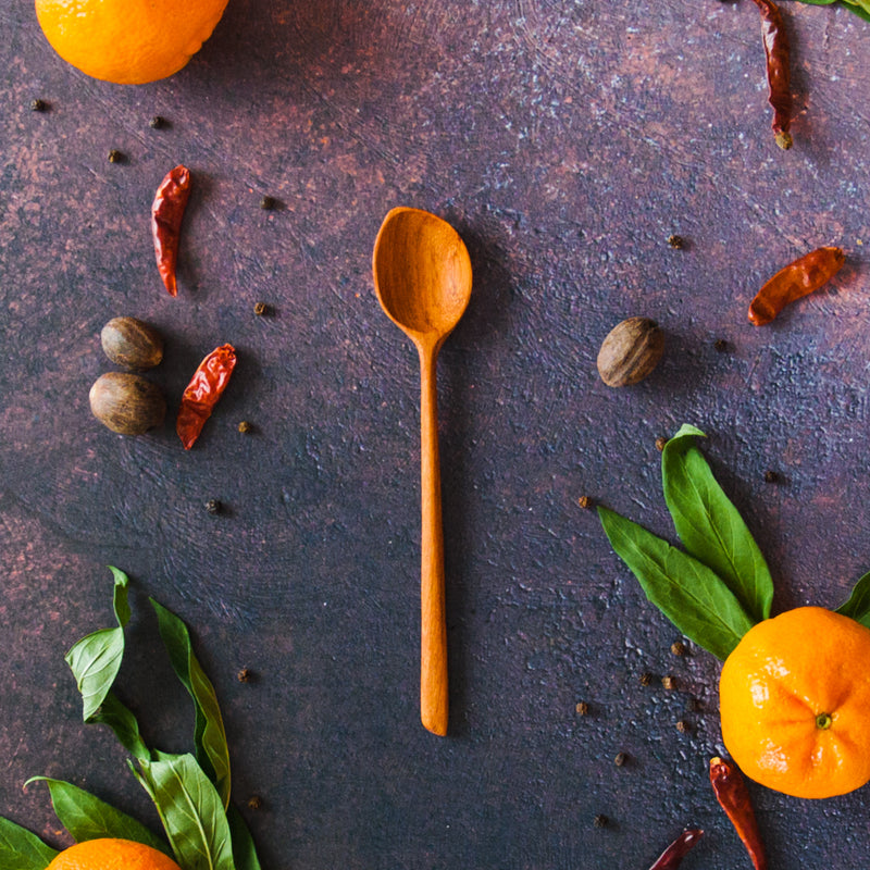 Carved wooden dip spoon on a dark background with oranges, greenery and red chili peppers