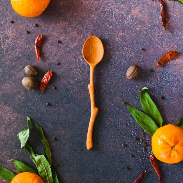 Wooden spoon with a twig shaped handle on a dark background with oranges, leaves and peppers