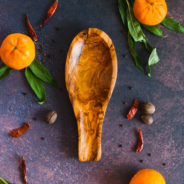 Olive wood spoon rest on a black rusty background with oranges, green leaves and red pepper surrounding it