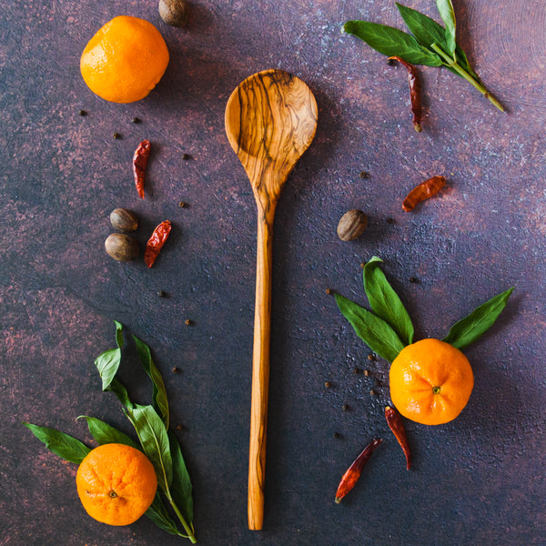 Long handled wooden spoon with a round head on a dark surface with clementines, greenery and red chilis