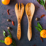 Set of two olive wood salad servers on a rusty dark surface surrounded by clementines, green leaves and red peppers