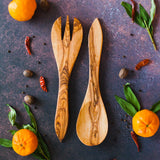 Large olive wood serving fork and serving spoon paired as salad servers on a dark background with oranges, leaves and chilis