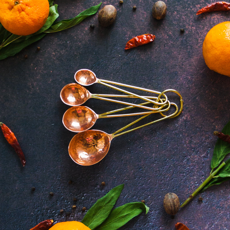 Copper and brass measuring spoons on a dark background with oranges, leaves and chilis surrounding it