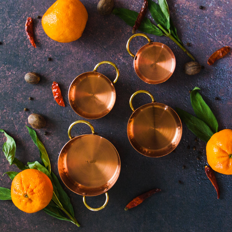 Four copper measuring cups of varying sizes on a dark background with oranges, green leaves and red chili peppers