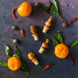 Olive wood wine bottle stoppers of four different shapes on a dark rusty surface with oranges, greenery and red peppers