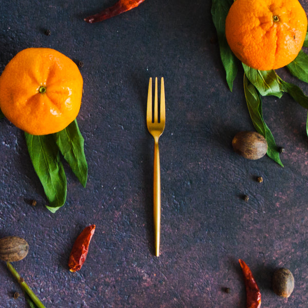 Small gold appetizer fork with three tines on a rusty black background with oranges and greenery