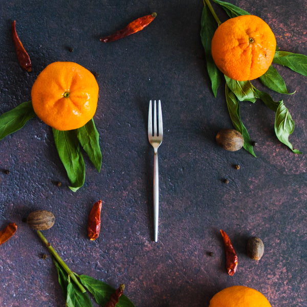 Silver appetizer fork on a dark rusty surface with clementines, green leaves and red peppers