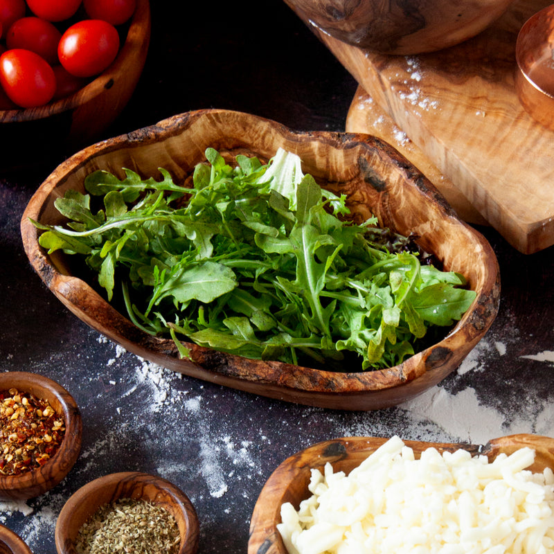 Medium wood dish with raw edges filled with arugula, on a dark background surrounded by pizza ingredients