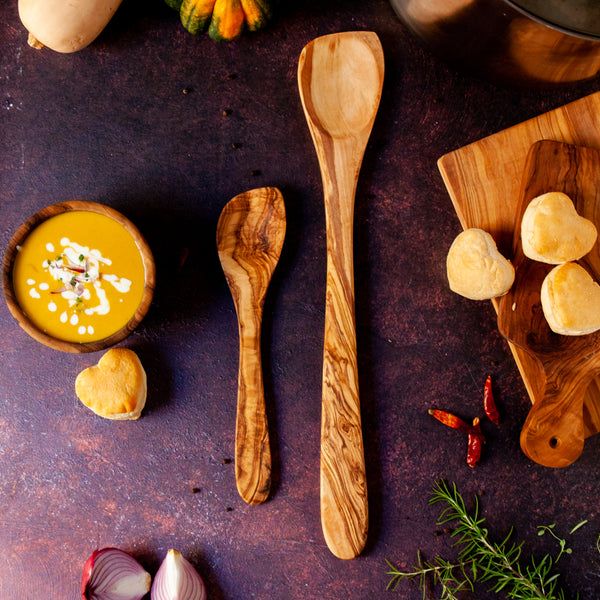 Pointed wooded spoon with extra long handle and similar smaller spoon on dark background with butternut squash soup and heart shaped biscuits