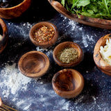 Four wooden pinch bowls on a dark background. Two of the bowls are filled with spices. They are all surrounded by pizza ingredients and are on a dark background with a dusting of flour.