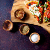 Four pinch bowls with seasonings, with a wooden board with pizza on it. All on a dark background