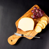 Silver cheese knife on wood paddle board with cheese, crackers and grapes on black